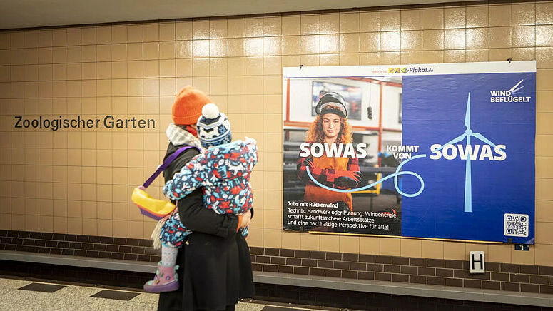 Mutter trägt Kind auf dem Arm und betrachten ein Plakat vom der Kampagne „Wind beflügelt“ in einer U-Bahnstation