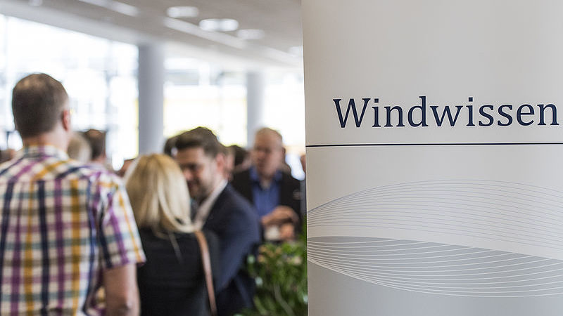 Ein Aufsteller mit dem Wort "Windwissen" ist zu sehen im Hintergrund sind Menschen die sich austauschen